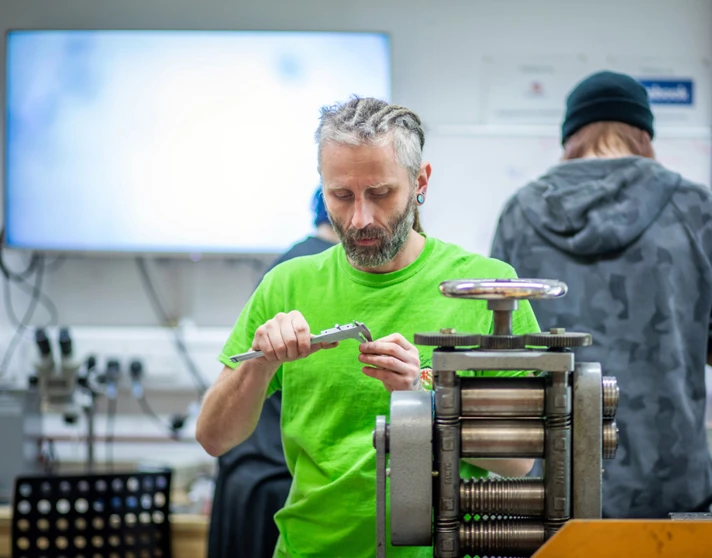 A male craftsman with braided hair and a beard, wearing a bright green shirt, measuring a piece of jewellery with calipers in a workshop filled with equipment. A male craftsman with braided hair and a beard, wearing a bright green shirt, measuring a piece of jewellery with calipers in a workshop filled with equipment.